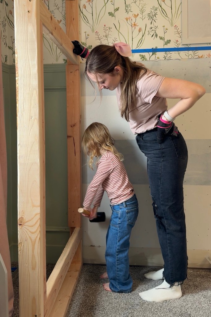 A child and an adult are collaborating on building custom twin over twin bunk beds, with the child using a mallet to tap a wooden frame. The room features floral wallpaper and partially completed walls.
