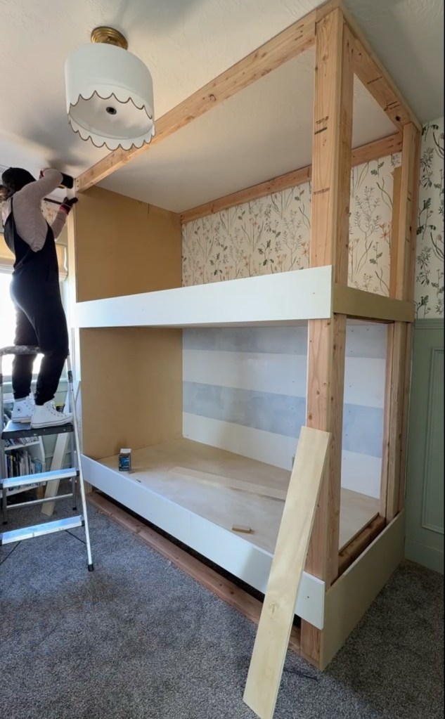 A person standing on a ladder while working on a partially built twin over twin bunk bed in a cozy room with floral wallpaper and a scalloped semi flush light.
