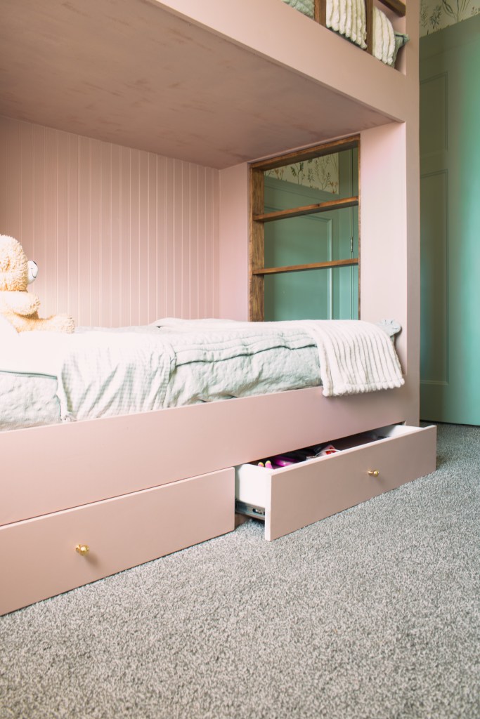 A close-up view of a pink twin over twin bunk bed with a cozy blanket, featuring storage drawers underneath, set against beadboard.