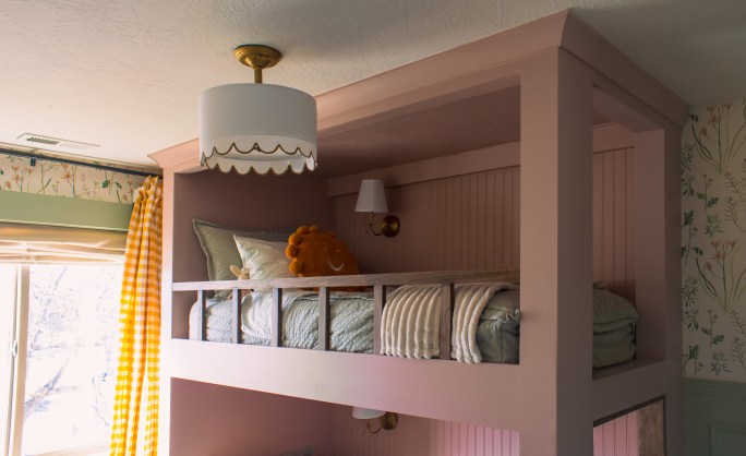 Built-in twin over twin bunk beds painted in a cozy pink color with a plush orange toy on the upper bunk, surrounded by a softly lit room with floral wallpaper.