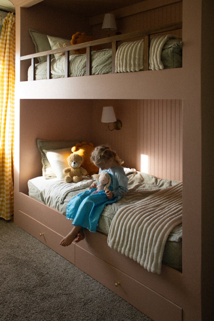 A child sitting on a lower bunk bed, dressed in blue, holding a doll, surrounded by stuffed animals, in a cozy and stylish room with custom pink built-in twin bunk beds. Yellow gingham curtains and beadboard, with sconces.