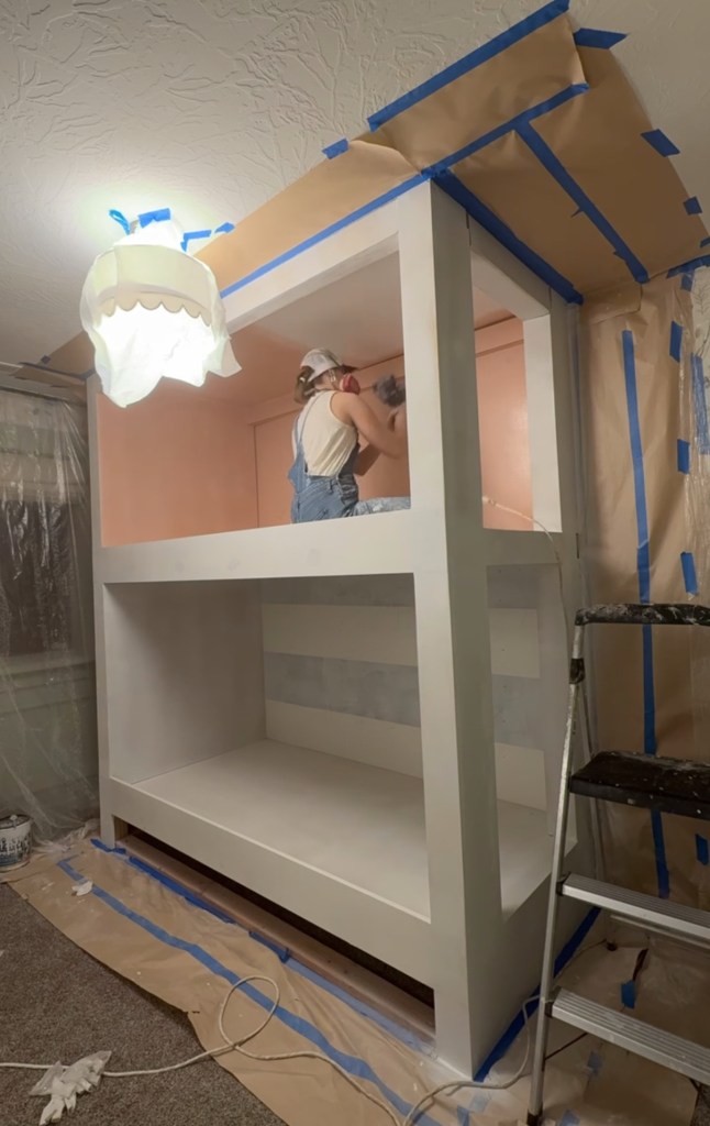 A person is painting a built-in twin over twin bunk bed in a pink color in a room with a protective tarp on the floor and painter's tape on the walls.