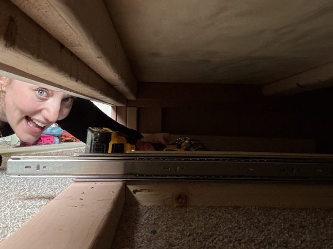 A person smiling while working on a DIY project under a bunk bed, with tools and drawer slides visible.
