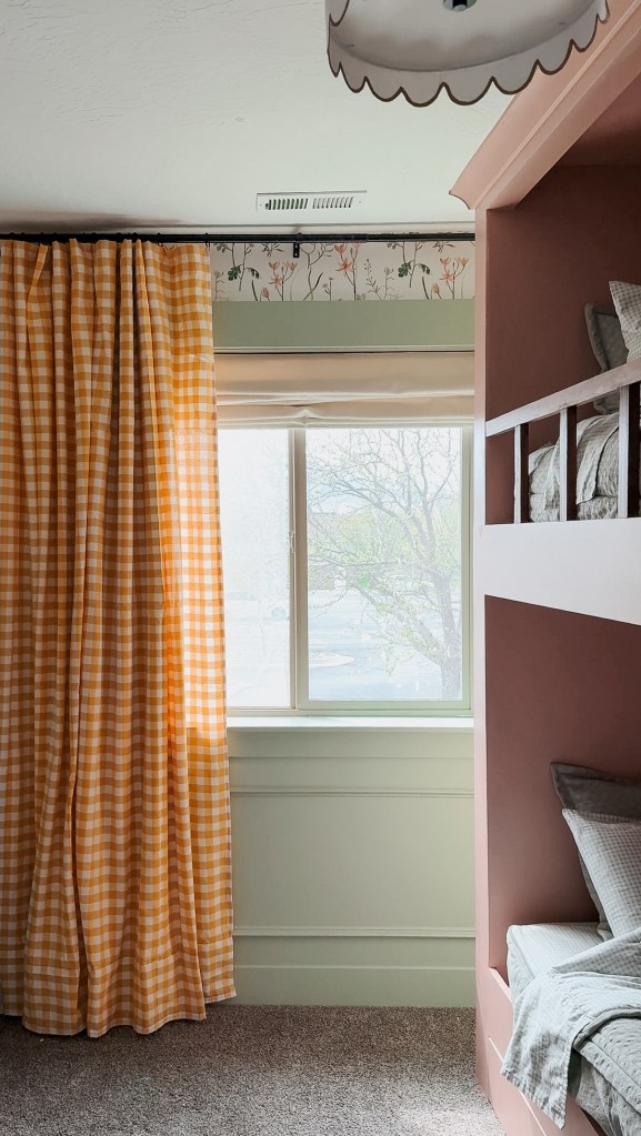 A cozy child's room featuring built-in twin bunk beds painted in a soft pink color, with a window dressed in yellow gingham curtains and green wall accents.
