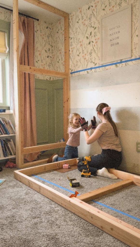 A mother and daughter are working together on building a twin over twin bunk bed frame, with the daughter wearing gloves and assisting. The room features floral wallpaper, a bookshelf, and a playful atmosphere.
